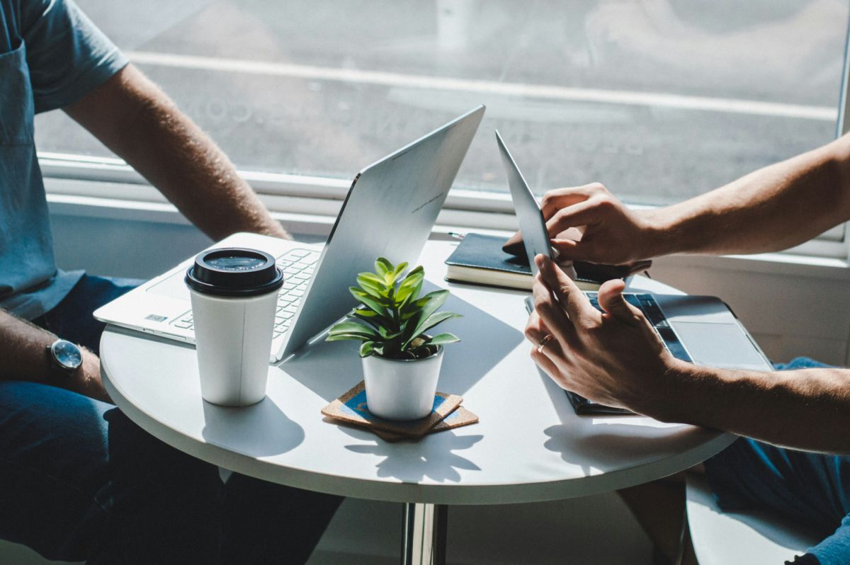 A photo of a two people having a meeting. A table with two laptops, a coffee cup and a plant